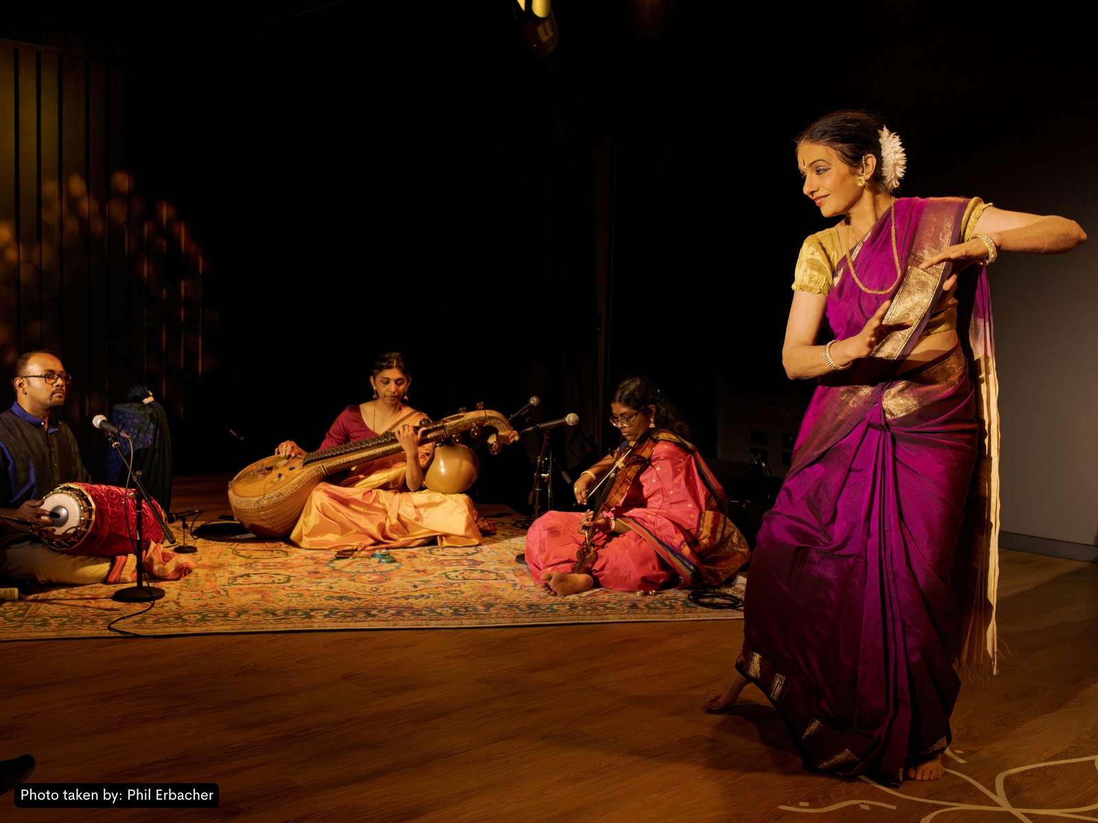 Wide-stage production photo — Bharatanatyam formation, deep amber/magenta lighting, immersive audience-close staging.